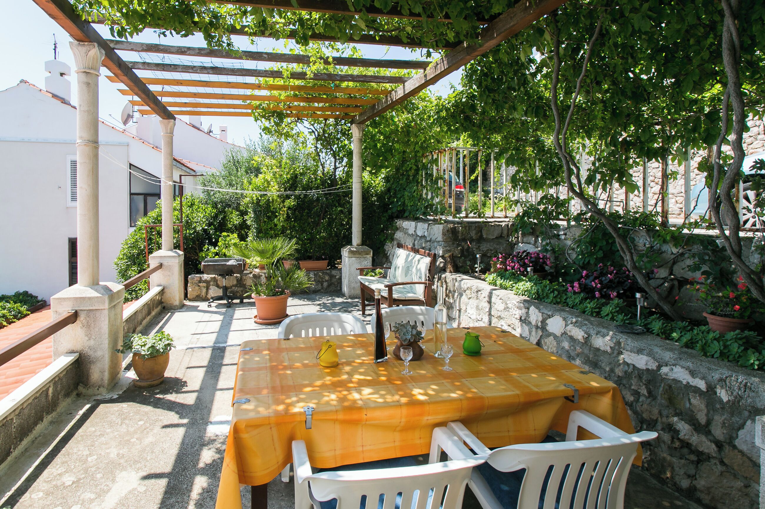 Une terrasse extérieure avec une table et des chaises, entourée de plantes en pot et d’un mur de pierre.