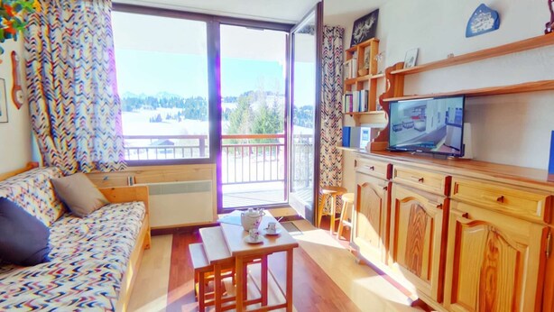 Ein gemütliches Wohnzimmer mit einer Couch, einem Esstisch und einem Fernseher. Ein Balkon bietet Blick ins Freie.