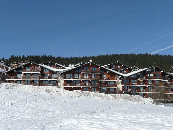 A row of multi-story wooden cabins with balconies, set against a snowy landscape and forested background.