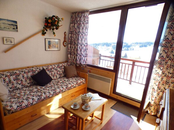A cozy room with a wooden sofa, a small table with cups and a teapot, a radiator, and a window with a view of a snowy landscape. - 2-room apartment for 4 in Les Saisies, comfort and stunning view (Les Saisies)