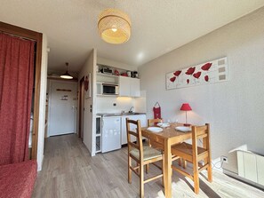 Une cuisine compacte avec des meubles blancs, un four à micro-ondes et un petit réfrigérateur. On y trouve une table à manger avec des chaises et une lampe rouge.
