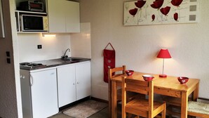 Une cuisine compacte avec des meubles blancs, un four à micro-ondes, une cuisinière et un évier. On y trouve une table en bois avec des chaises et une lampe rouge.