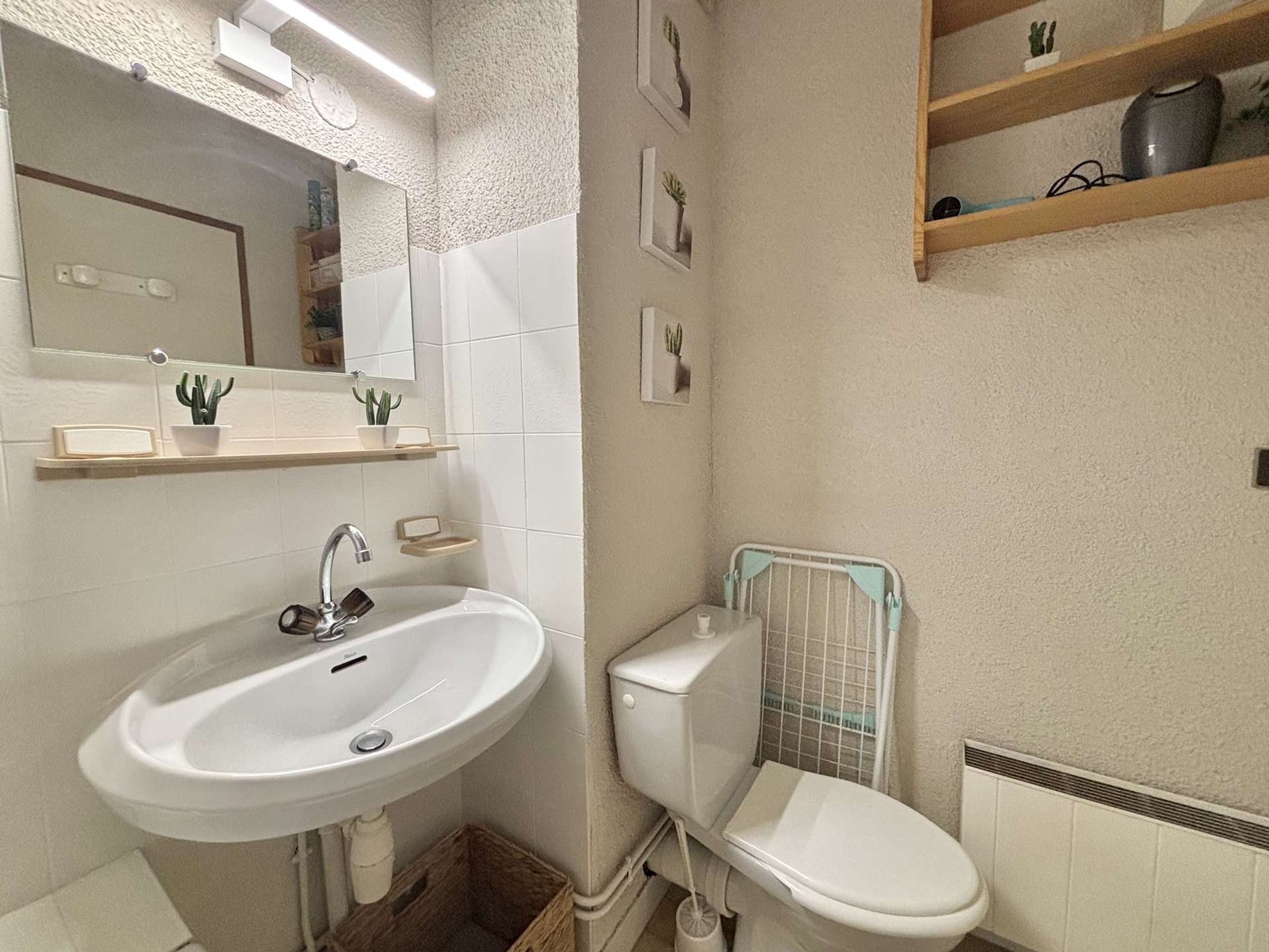 Un baño con un lavamanos blanco, espejo, inodoro y un estante con pequeñas plantas.