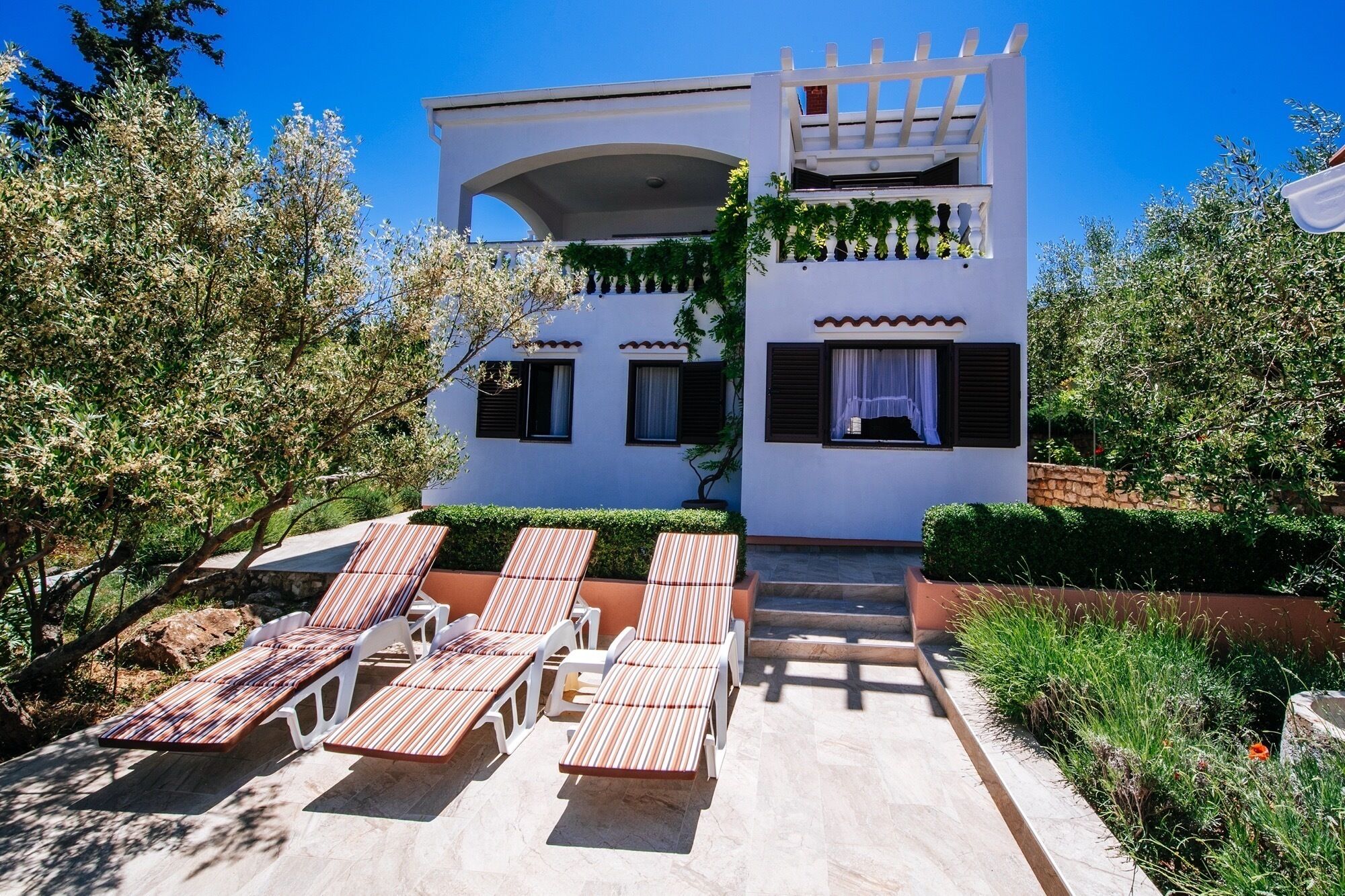 A two-story building with a white facade, a covered outdoor seating area with striped lounge chairs, and a stone pathway leading to the entrance.