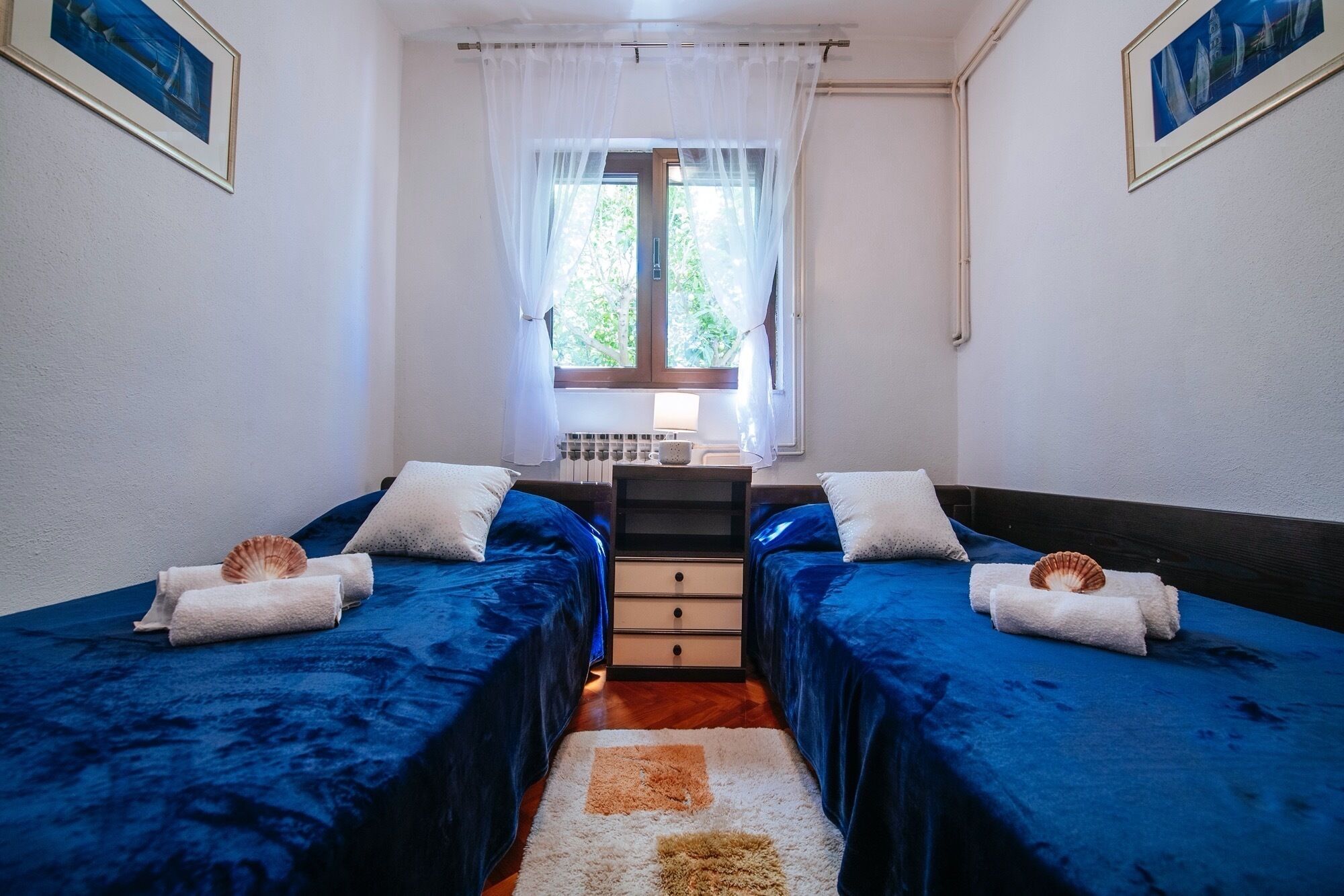 A room with two beds, each with a blue bedspread and a white pillow, a small table with a lamp, and a window with sheer curtains.