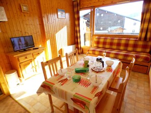 A wooden interior with a dining table set for two, a television on a wooden stand, and a window with a view of buildings outside.