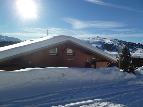 Una cabaña de madera con techo inclinado, rodeada de montañas nevadas y un cielo despejado.