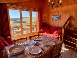 A dining area with a table set for two, wooden walls, a red sofa, and a view of a green landscape.