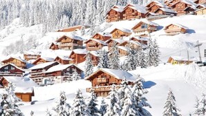 Un village de montagne enneigé, avec des chalets en bois, entouré d’arbres chargés de neige et sous un ciel dégagé.