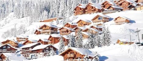 A snow-covered mountain village with wooden chalets, surrounded by snow-laden trees and a clear sky.