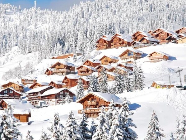 A snow-covered mountain village with wooden chalets, surrounded by snow-laden trees and a clear sky.
