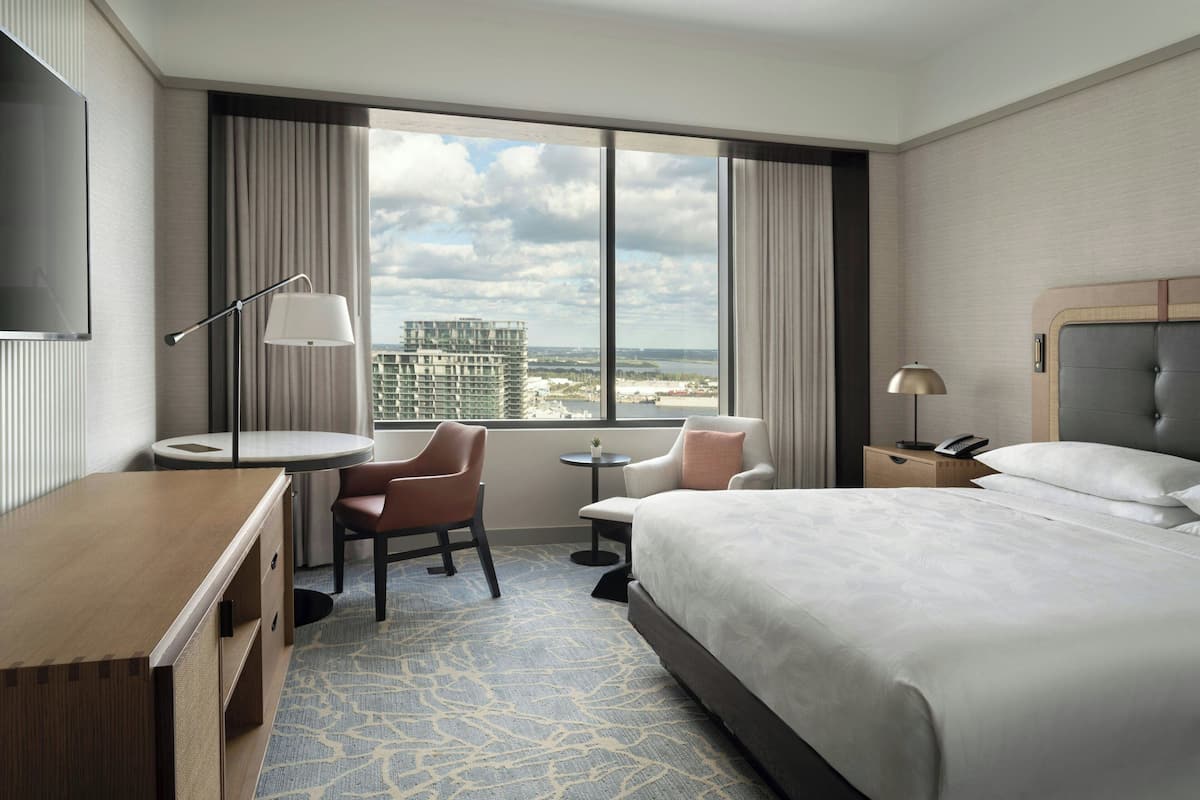 A hotel room with a large bed, a desk, a chair, a small table, a TV, and a window with a city view.