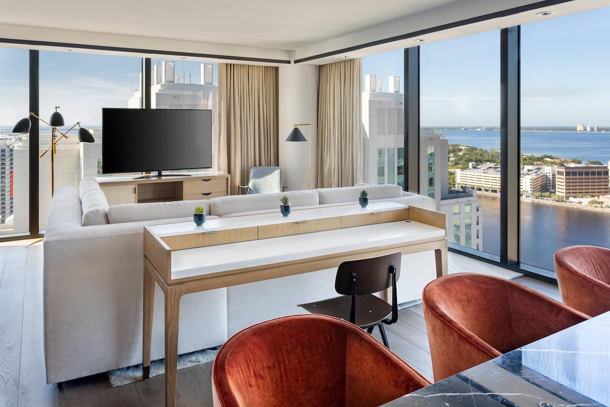 A modern hotel room with a large window, a flat-screen TV, a sofa, a dining table, and two chairs.