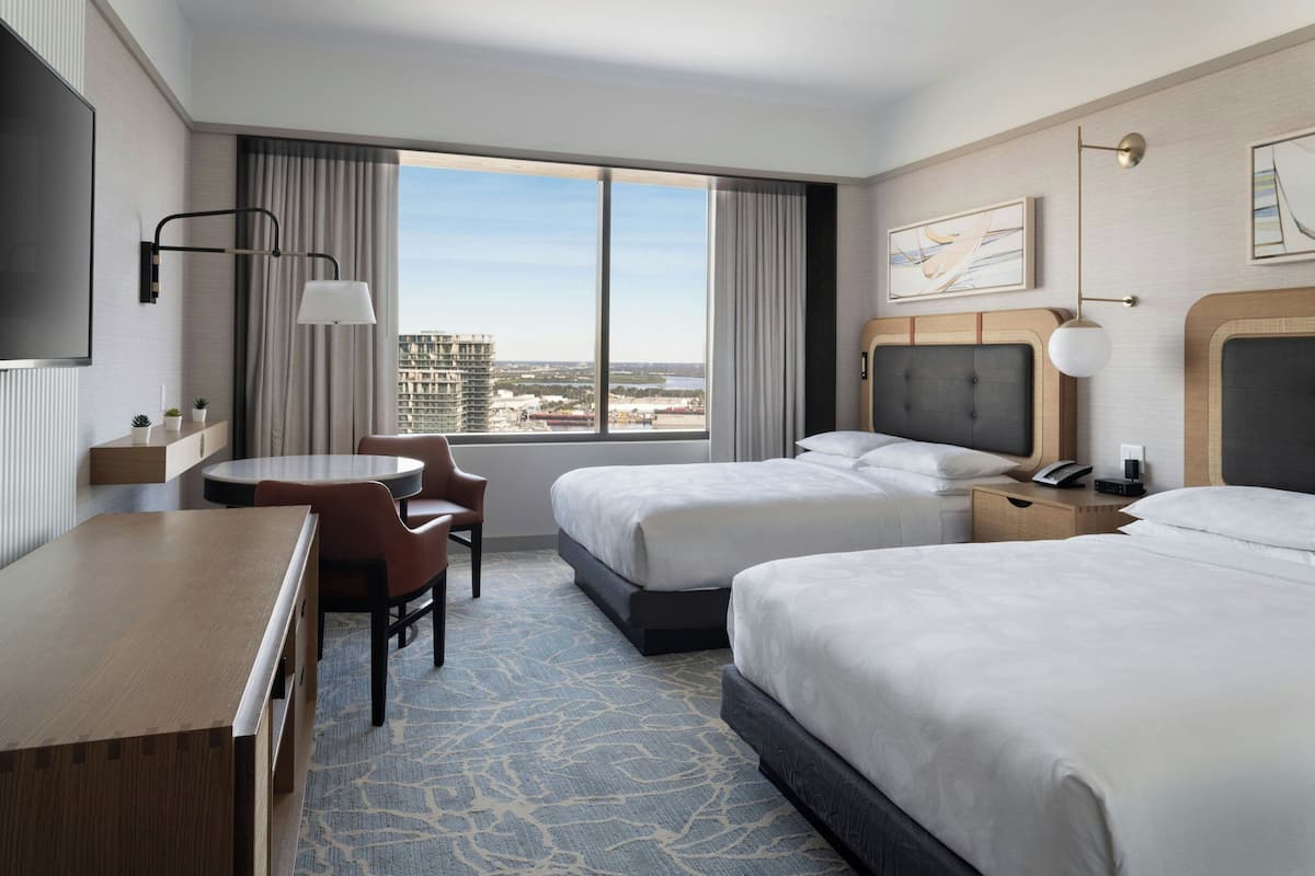 A hotel room with two beds, a desk, a chair, a TV, and a large window with a city view.