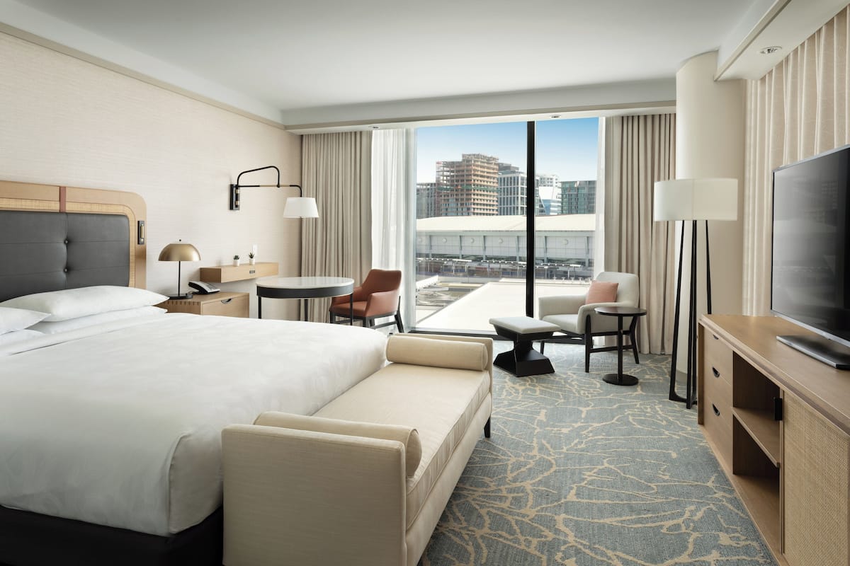 A hotel room with a large bed, a desk, a chair, a TV, and a view of the city through the window.