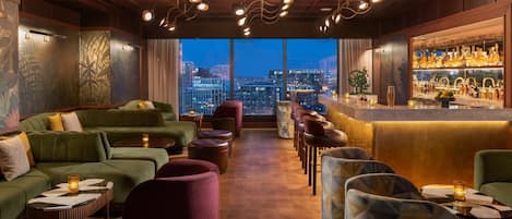 A modern lounge with green sofas, red and blue chairs, a bar area with bottles, and a city view through large windows.