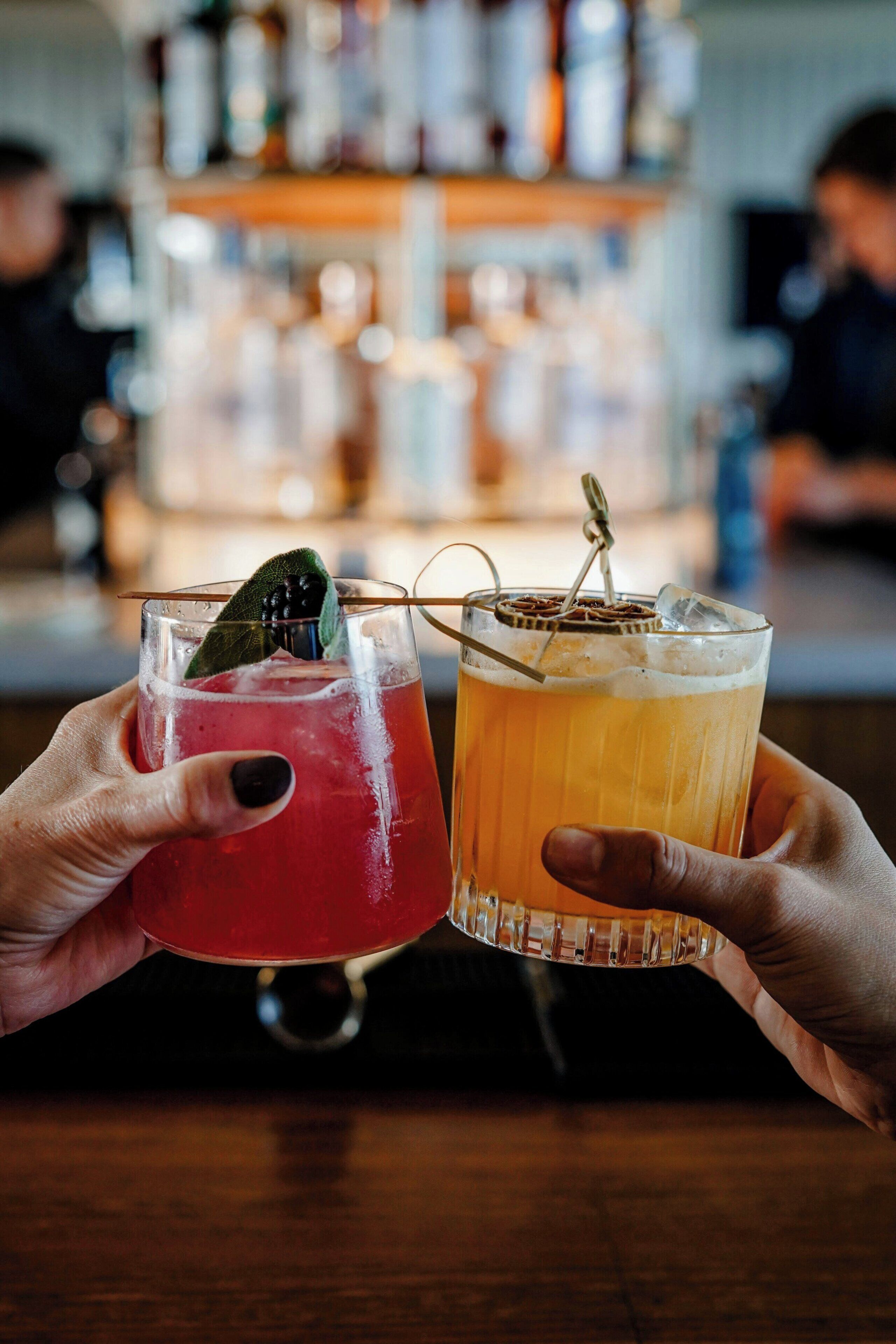Zwei Hände halten Gläser mit farblich unterschiedlichen Getränken, eines mit Limettenscheibe, an einer Bar mit Flaschenregalen im Hintergrund.
