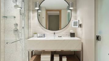 Une salle de bain moderne avec une douche à parois de verre, un miroir rond, un lavabo avec un plan de toilette en marbre et une étagère en bois où sont posées des serviettes.