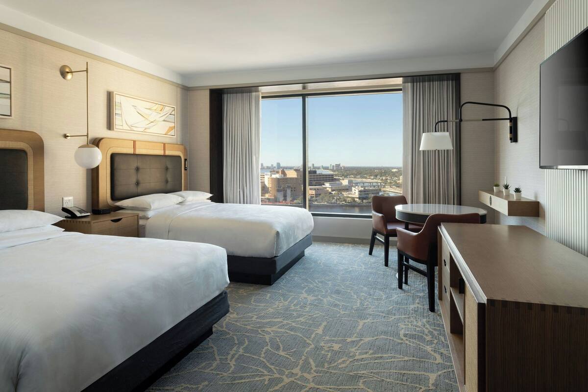 A hotel room with two beds, a desk, a chair, a TV, and a large window offering a city view.