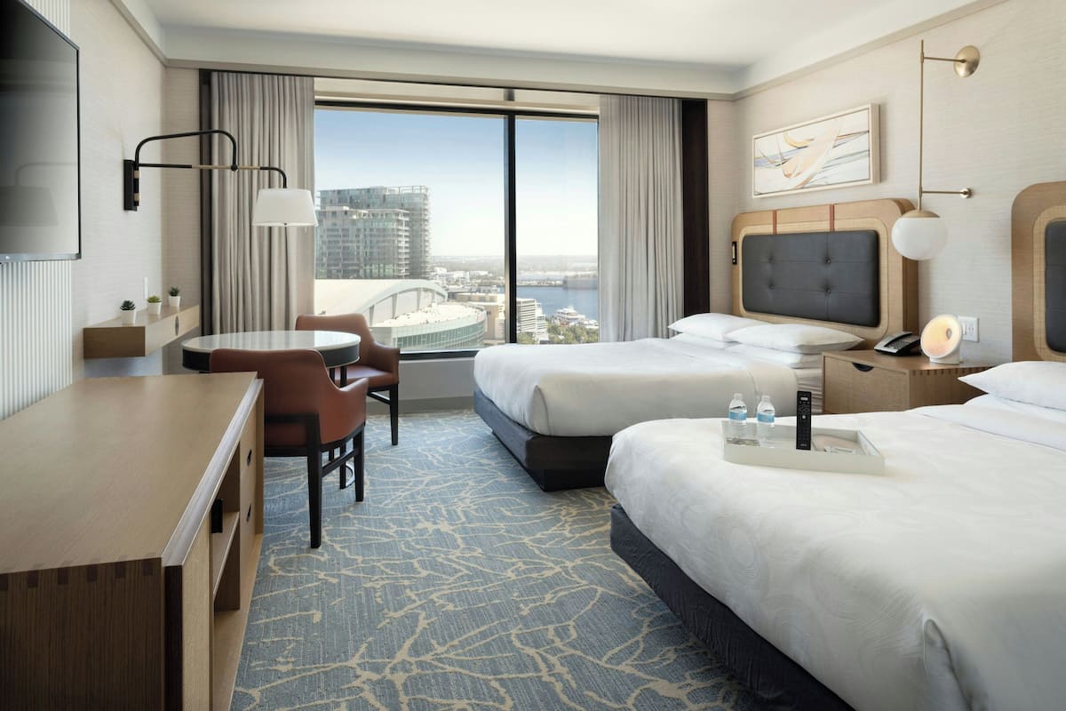 A hotel room with two beds, a desk, a chair, a TV, and a large window with a city view.