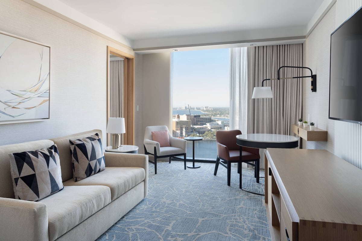 A hotel room with a sofa, armchairs, a small table, a TV, and a view of the city.