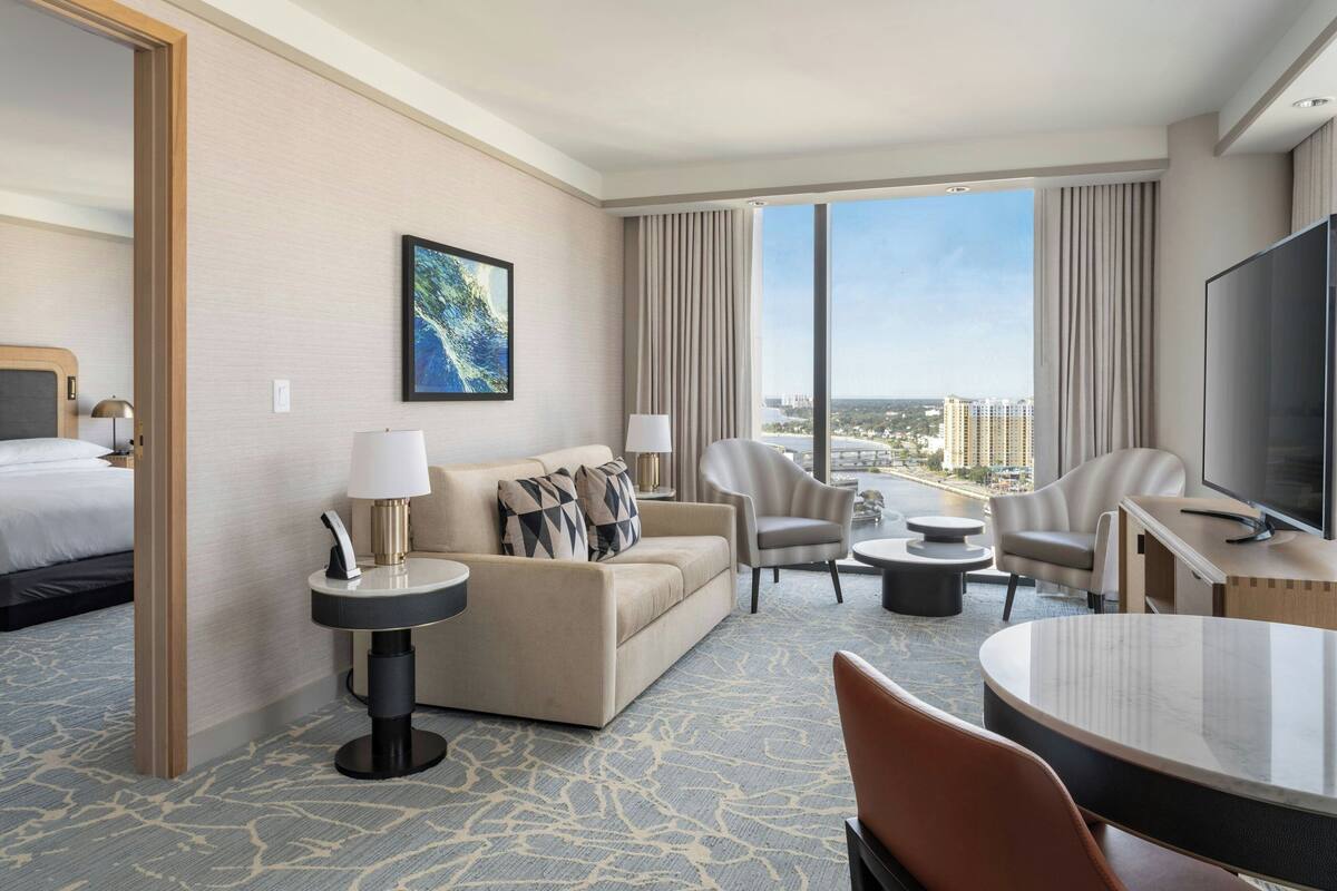 A hotel room with a bed, a sofa, two armchairs, a TV, and a large window offering a city view.