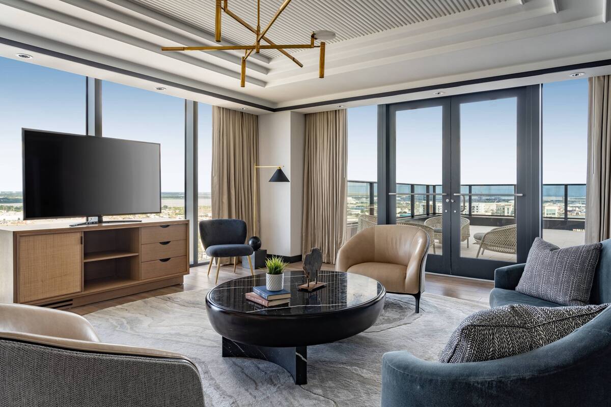 A modern hotel room with a large flat-screen TV, comfortable seating, a balcony with a view, and a stylish ceiling design.