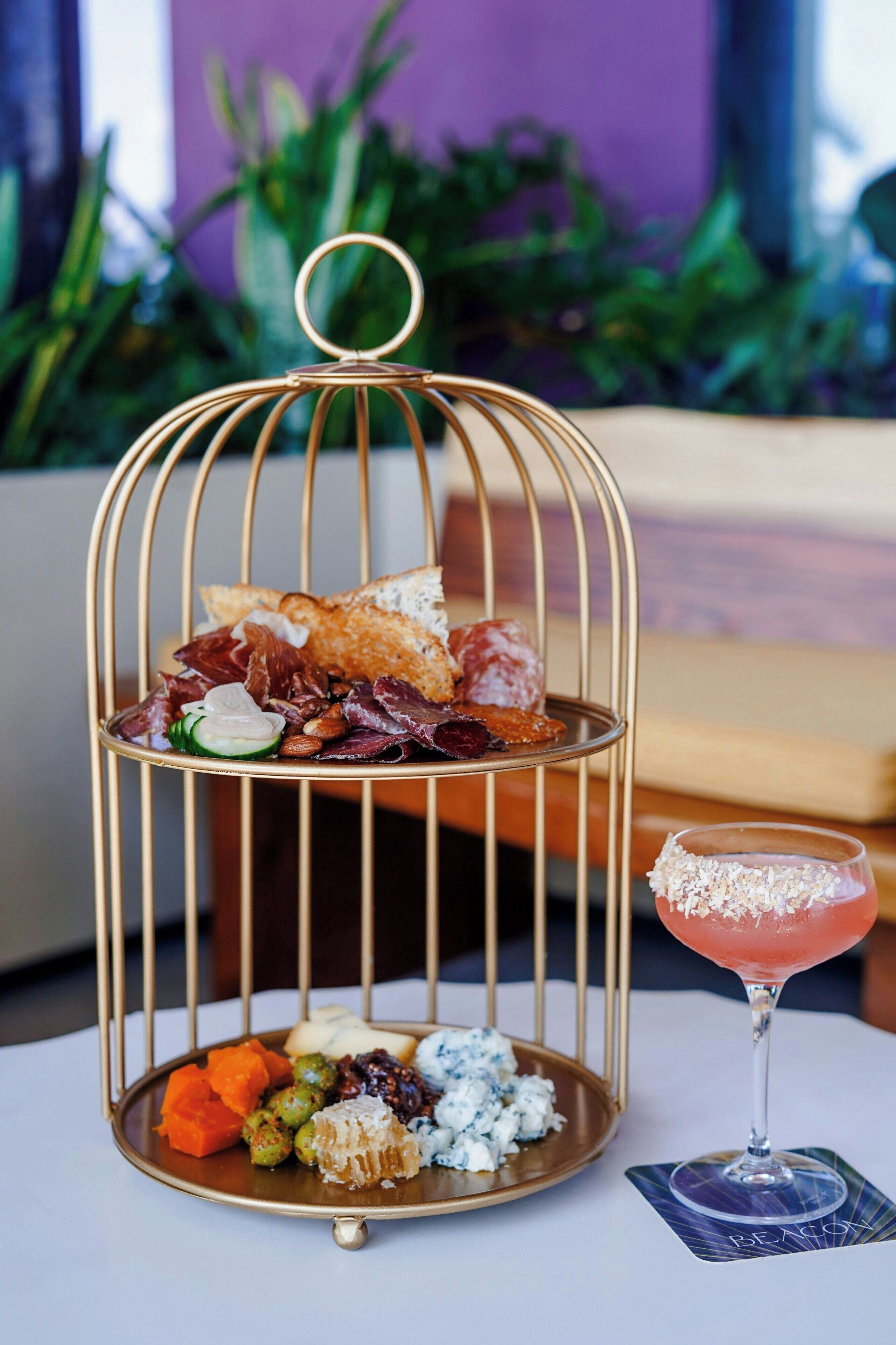 Un plateau de service en métal à deux niveaux, sur lequel sont disposés divers amuse-gueules et un cocktail, est posé sur une table.