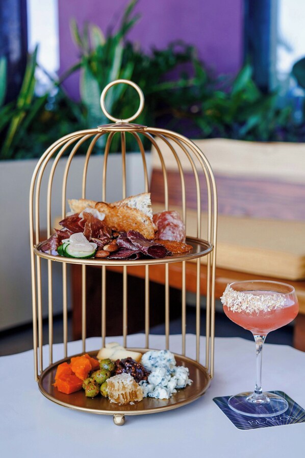 Una bandeja metálica de dos niveles con aperitivos variados y un cóctel sobre una mesa.