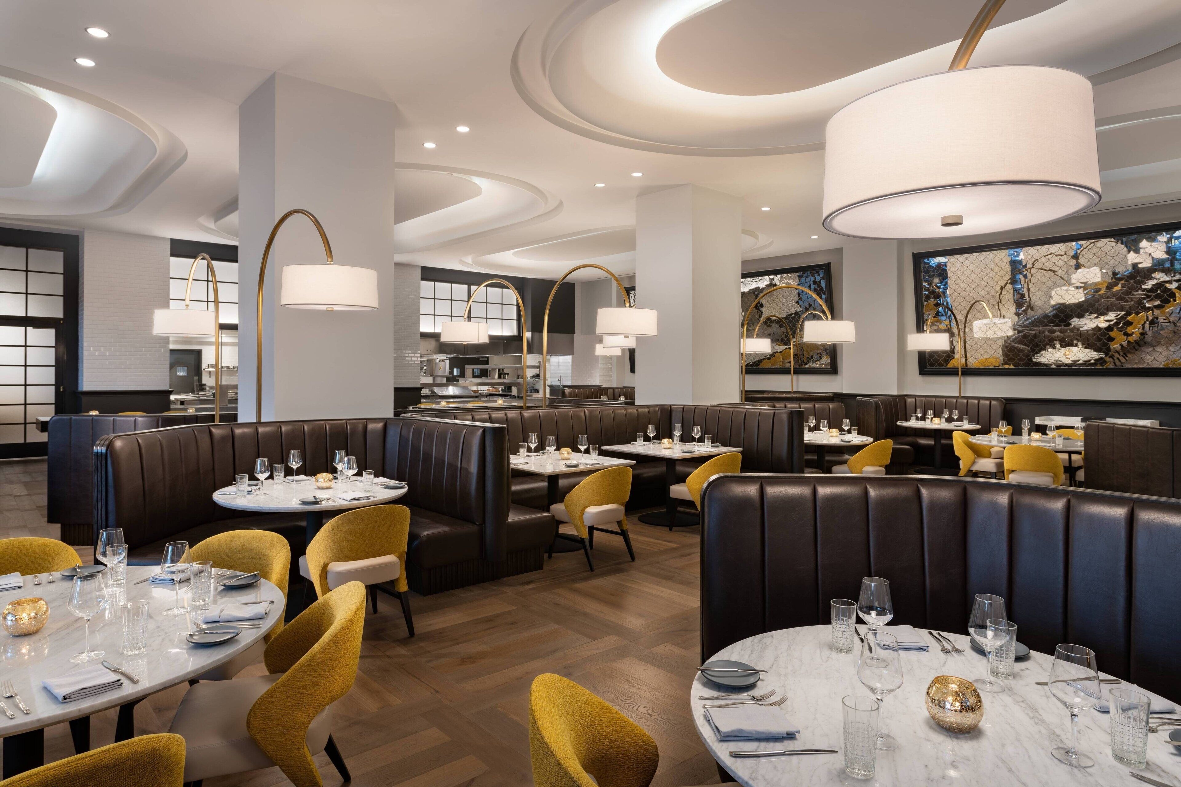 Interior moderno de restaurante con cabinas y mesas redondas, destacando un área central para comer con piso de mármol y sillas amarillas.