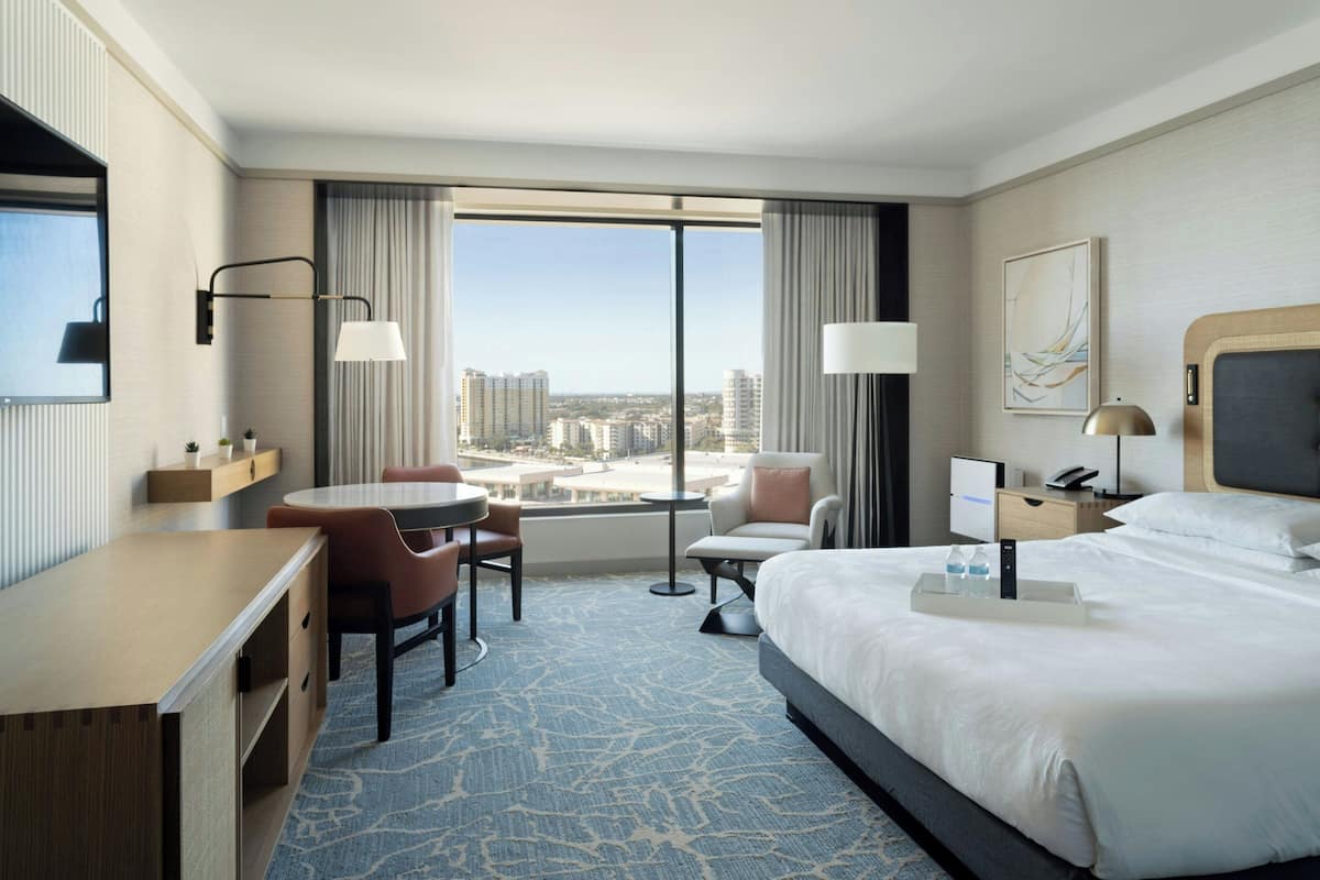 A hotel room with a large bed, a desk, a chair, a small table, and a view of the city through large windows.