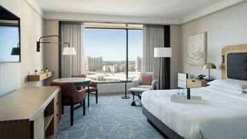 Ein Hotelzimmer mit einem großen Bett, einem Schreibtisch, einem Sessel, einem kleinen Tisch und Blick auf die Stadt durch große Fenster.