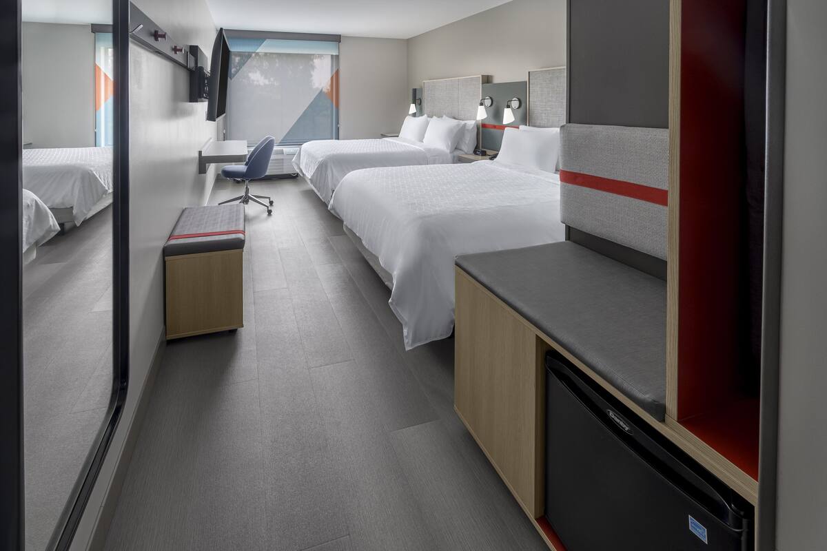 A hotel room with two beds, a desk, a chair, and a small kitchenette.