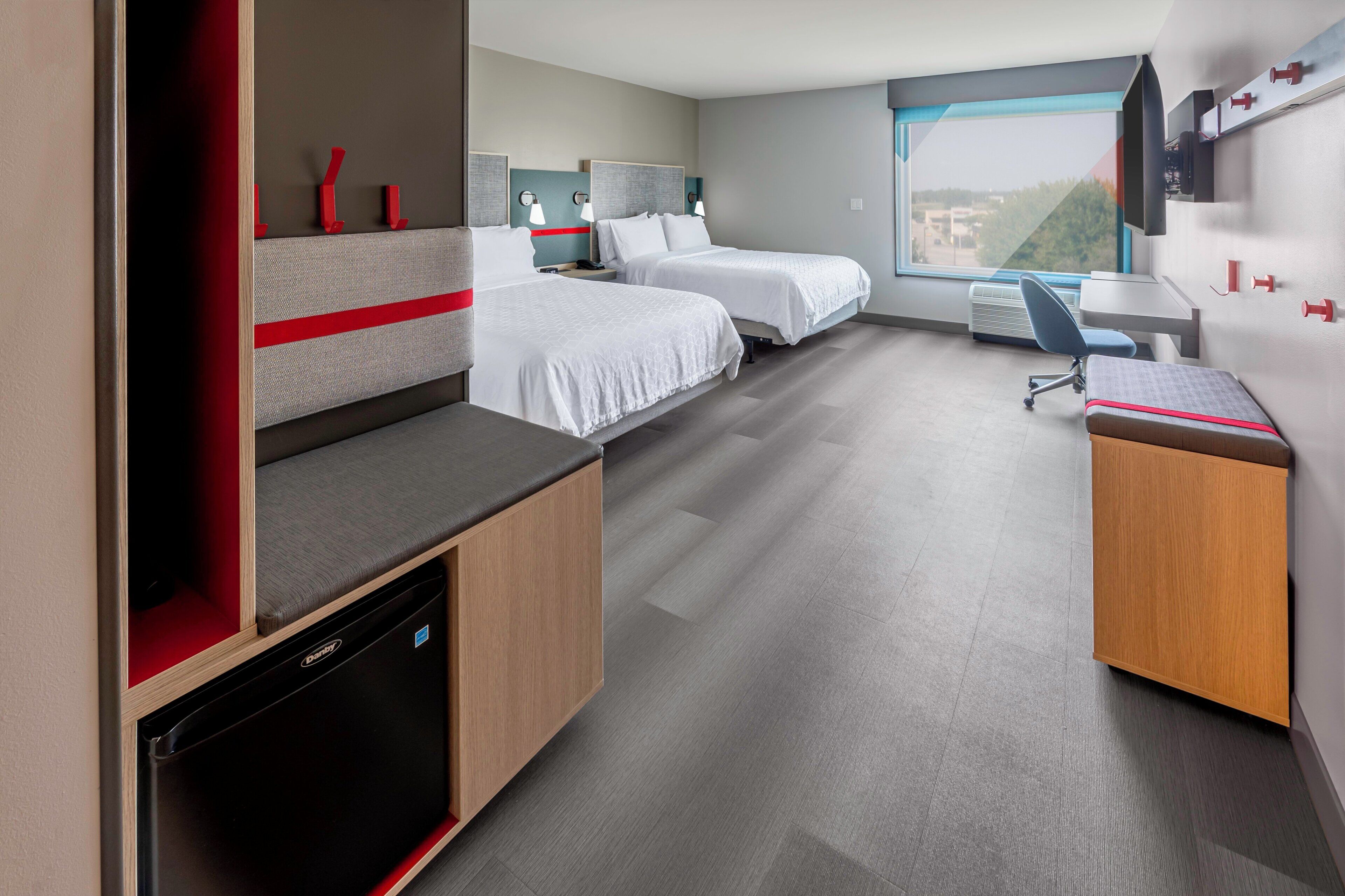 A hotel room with two beds, a desk, a chair, a built-in dishwasher, and a large window.