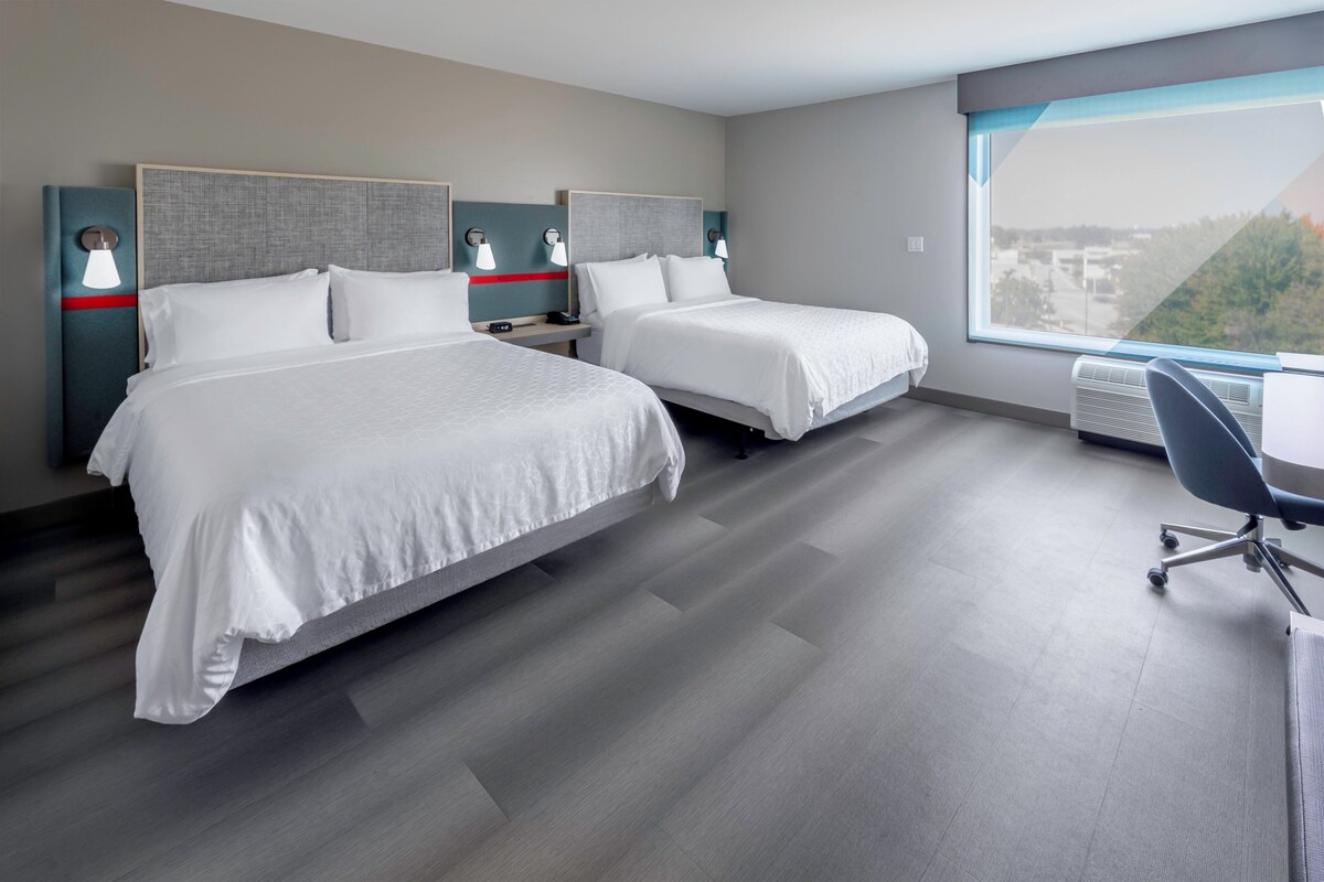Ein modernes Hotelzimmer mit zwei Betten, einem Schreibtisch und einem großen Fenster mit Blick auf die Stadt.