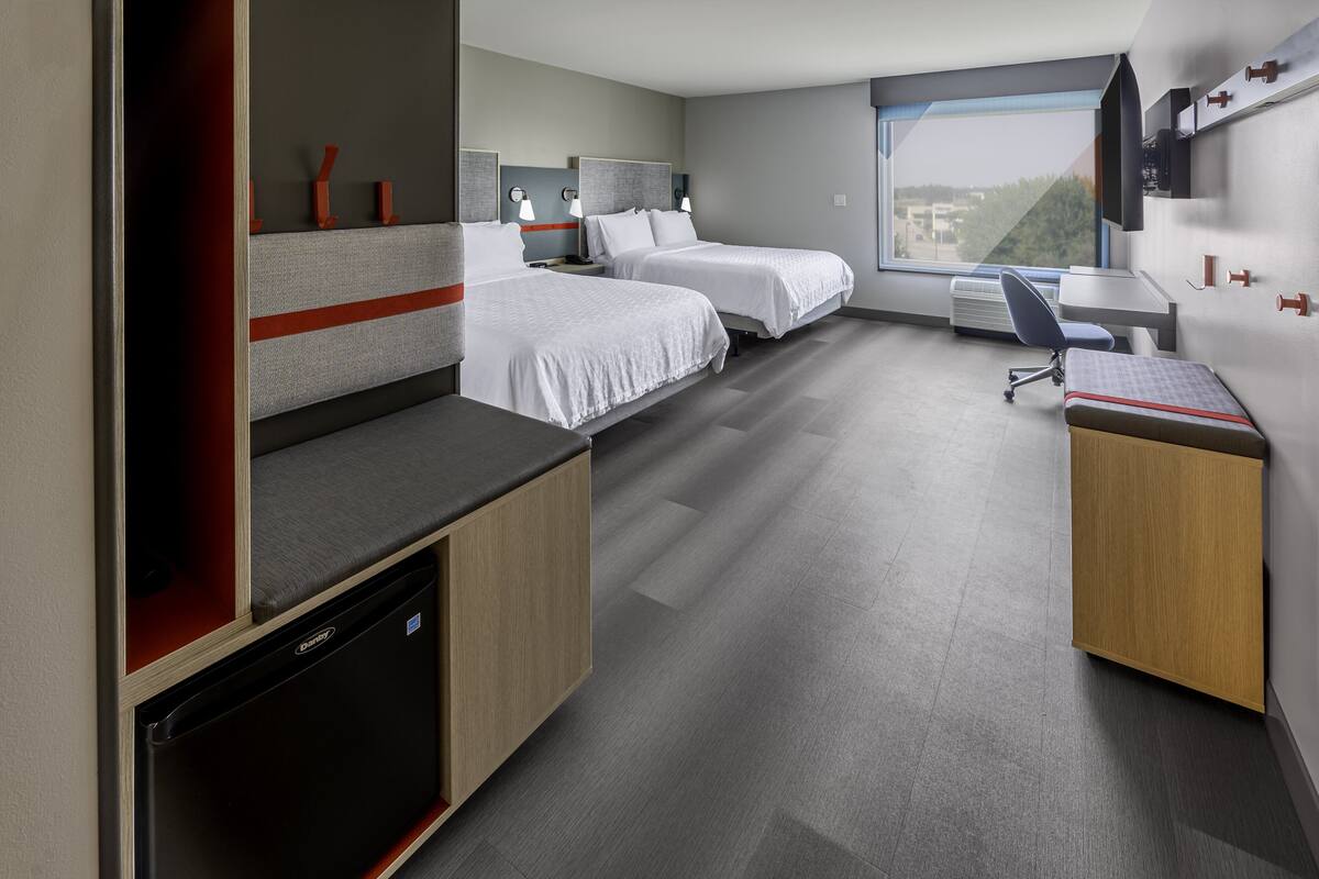 A modern hotel room with two single beds, a desk, a chair, a built-in mini-fridge, and a large window offering a city view.