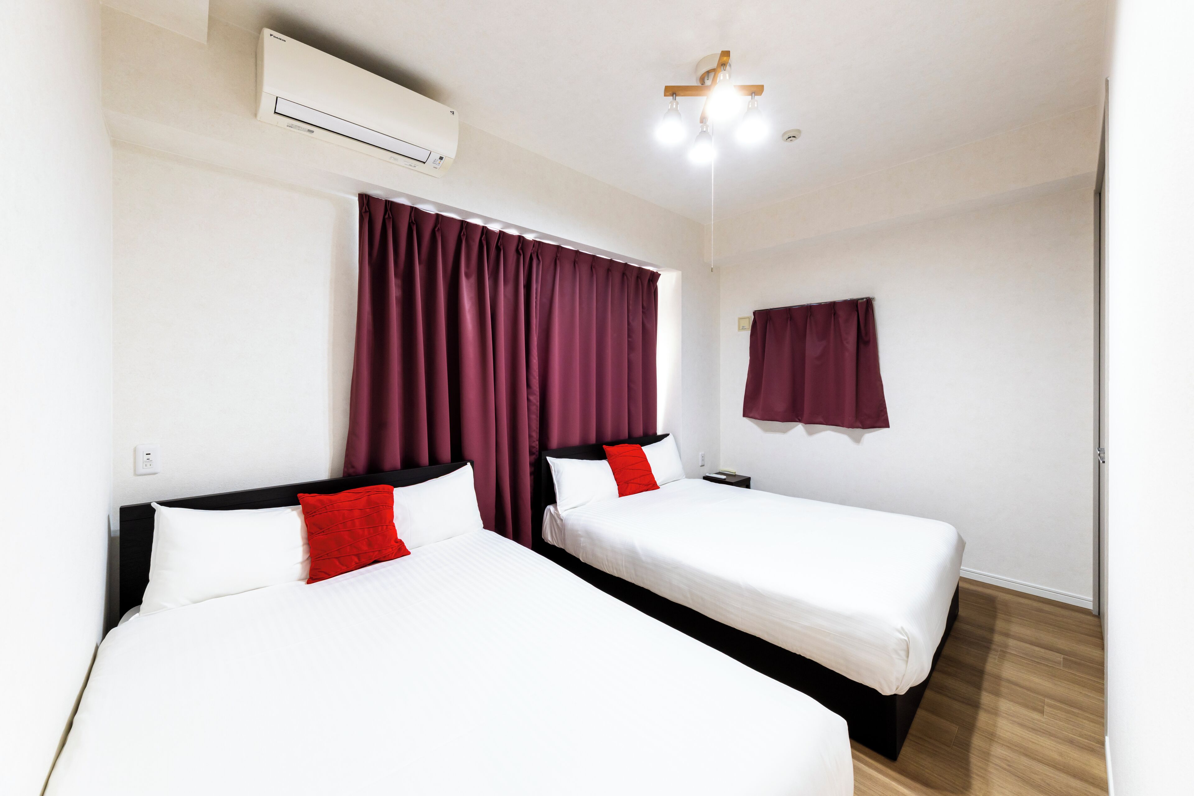 Una habitación de hotel con dos camas, una ventana con cortinas, un aire acondicionado, una mesita de noche y una lámpara fijada a la pared.
