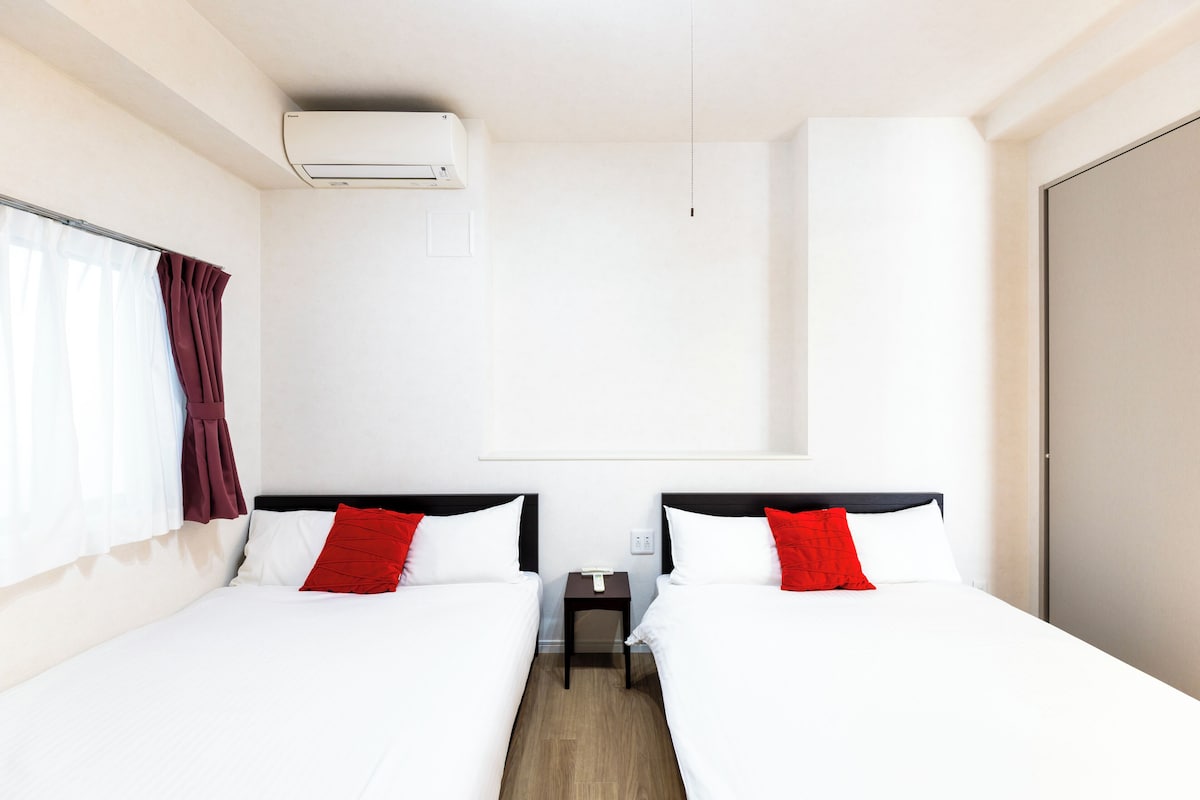 Une chambre d’hôtel avec deux lits, du linge de lit blanc, des oreillers rouges, une petite table de chevet et une unité de climatisation.