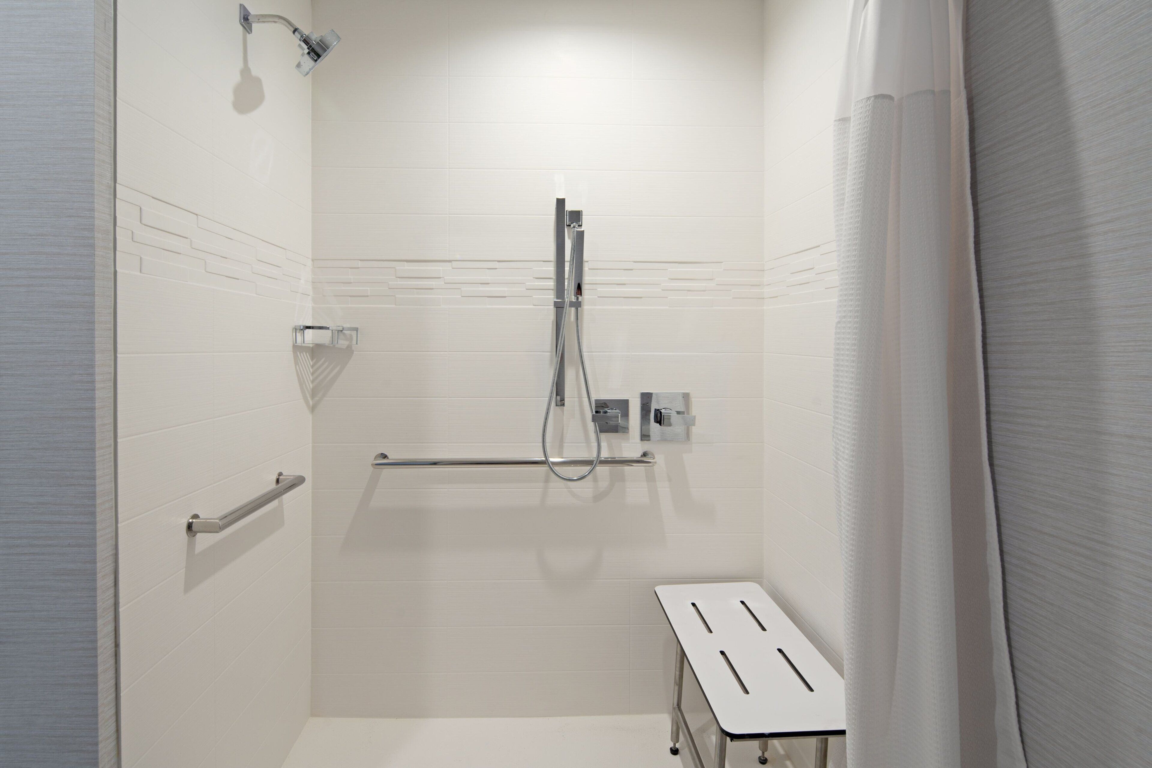 A modern bathroom with white tiles, a shower with a handheld showerhead, a towel rack, and a small table.