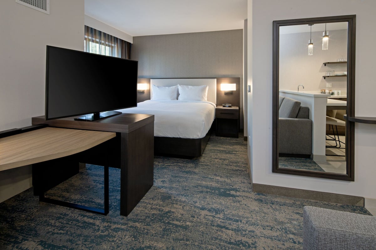 A hotel room with a bed, a TV, a desk, a mirror, and a small kitchenette.