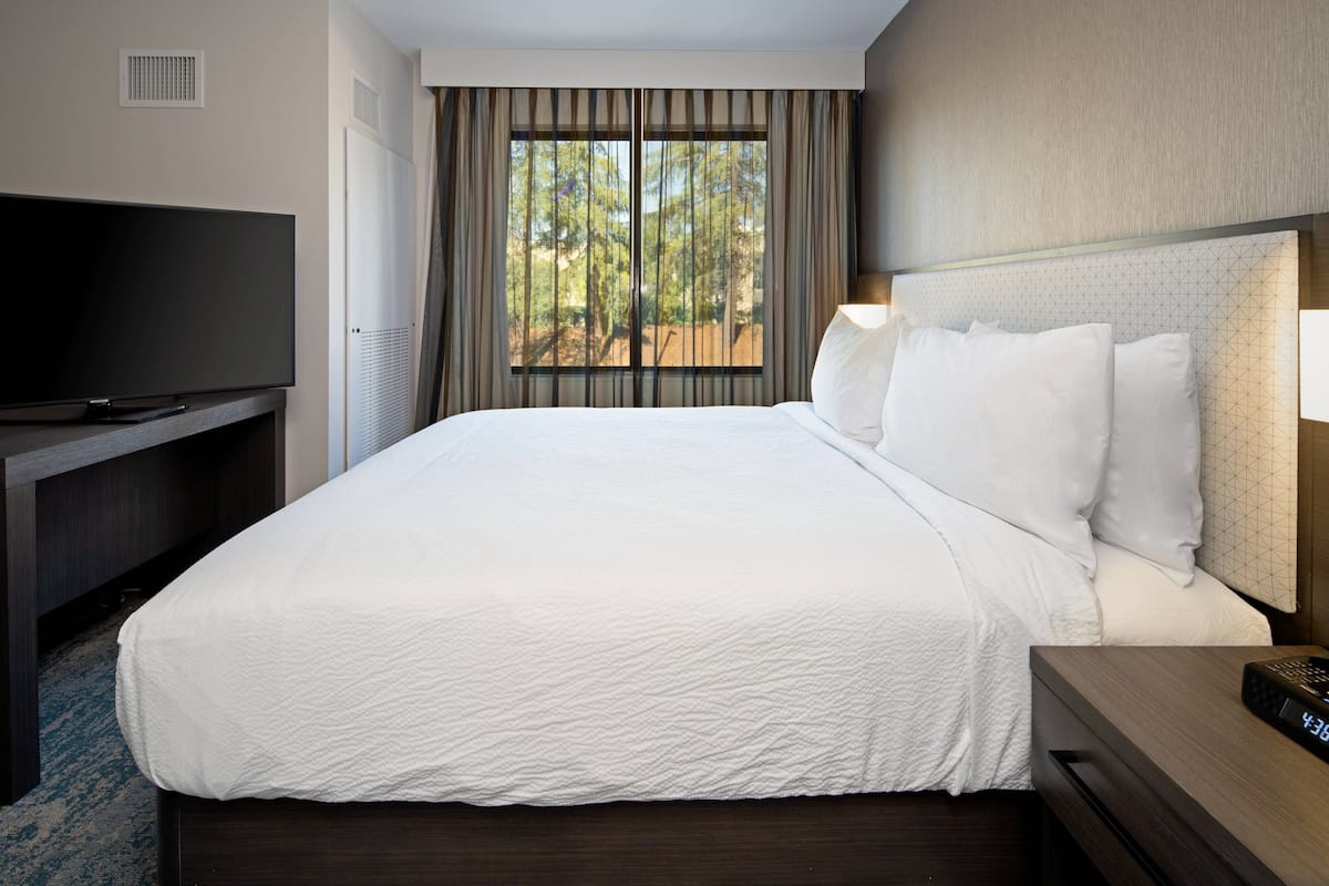 A hotel room with a large bed, a TV on a stand, a nightstand with a clock, and a window with curtains.