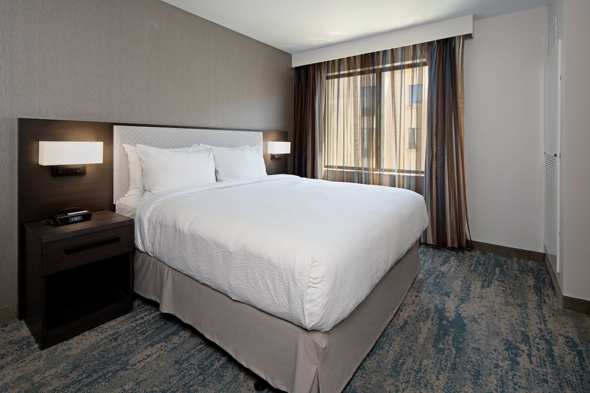 A hotel room with a large bed, a nightstand with a lamp, and a window with curtains.