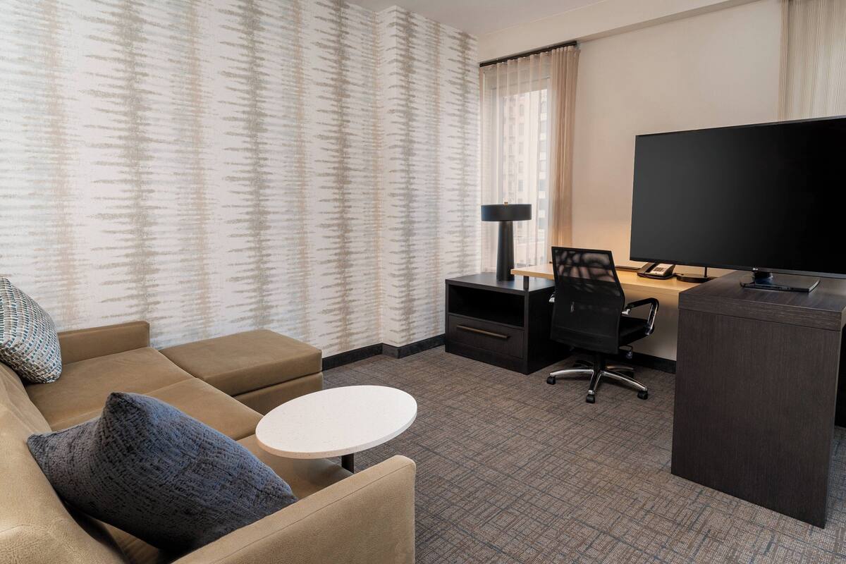 Hotelkamer met een bank, een klein rond tafeltje, een bureau met stoel, een televisie en een groot raam met gordijnen.