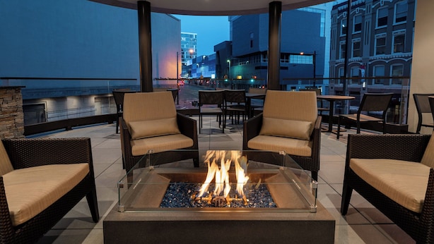 A modern outdoor seating area with a glass-top fire pit surrounded by wicker chairs, overlooking a cityscape at dusk. - Residence Inn by Marriott Grand Rapids Downtown (Grand Rapids)