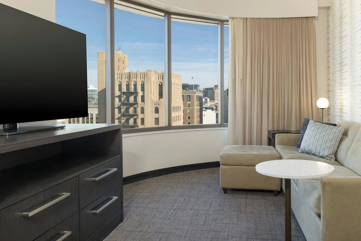 Hotelkamer met groot raam, flatscreen televisie, een bank, een klein rond tafeltje en een minibar.