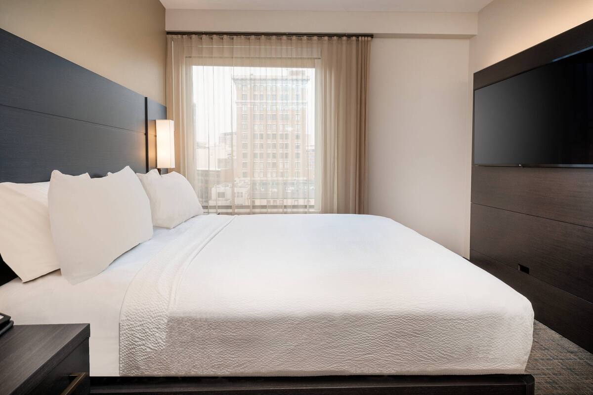Een hotelkamer met een groot bed, een flatscreen-tv, een raam met gordijnen en uitzicht op de stad.