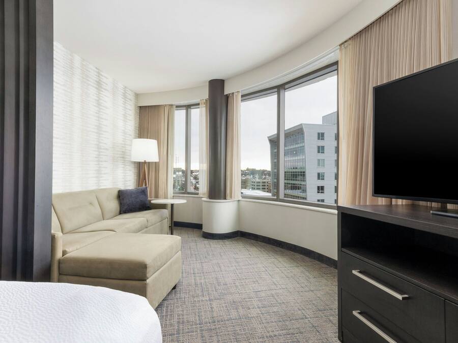 A hotel room with a large bed, a sofa, a TV, a small table, and a window offering a city view.