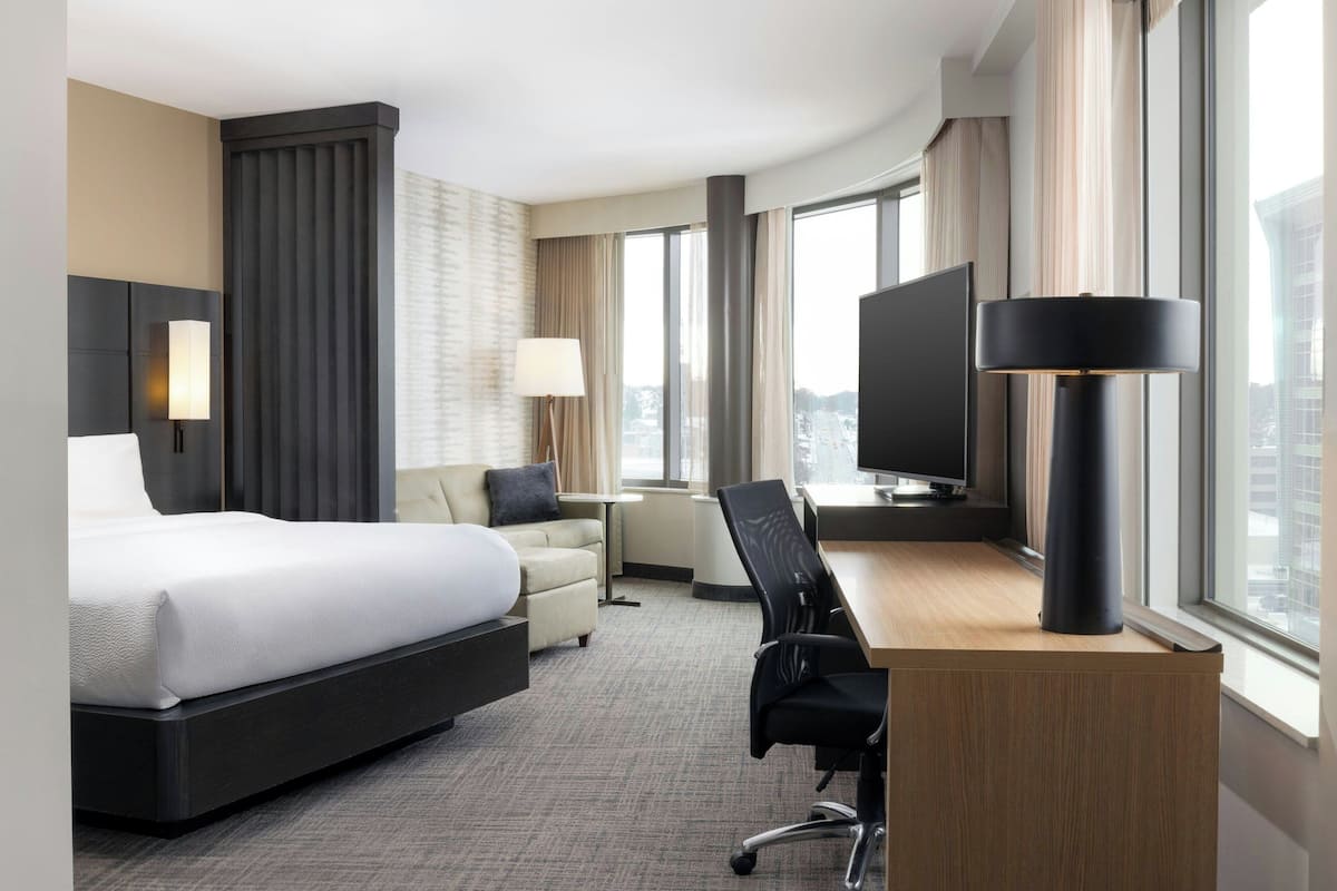 Een moderne hotelkamer met een groot bed, een bureau, een stoel, een televisie en een groot raam met uitzicht op de stad.