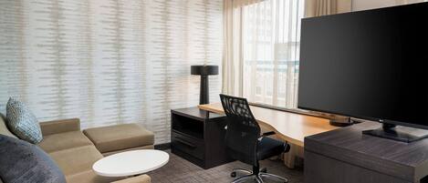 A hotel room with a sofa, a desk with a chair, a television, and a window with curtains.