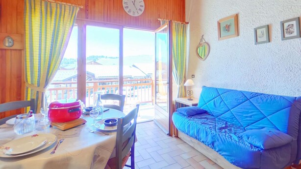 Um quarto aconchegante com um sofá azul, uma mesa de jantar posta com pratos e talheres, um relógio na parede e vista para um edifício pela janela.